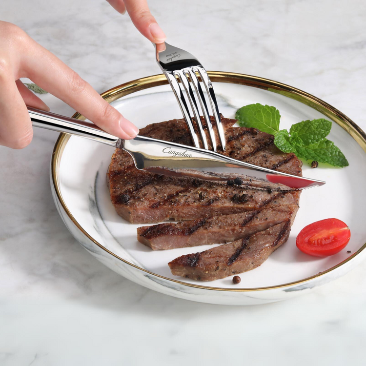 Hand holding a fork and steak knife cutting a grilled steak on a white plate with a cherry tomato and mint leaves.