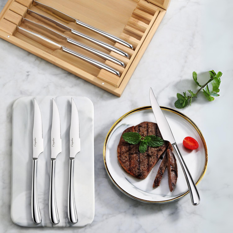 Eight stainless steel steak knives in a bamboo box, with three knives on a tray and one cutting a steak on a plate.