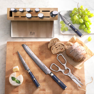 
                    
                      Wooden knife block with seven knives, two knives and a meat fork on a cutting board with bread, cheese, and grapes.
                    
                  