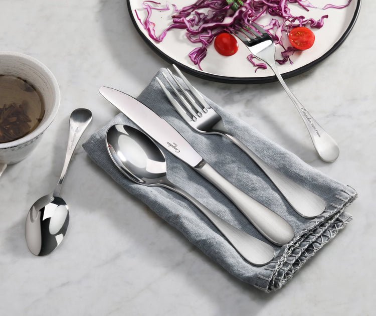 Stainless steel dinner fork with matte finish handle placed on a gray napkin alongside a knife and spoon on a marble surface.