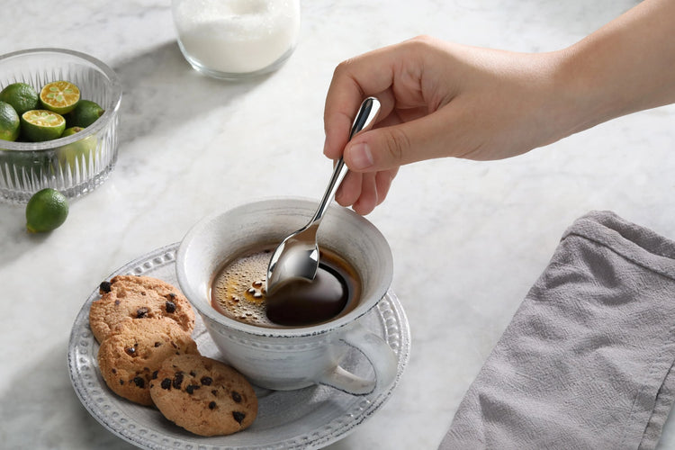 Hand holding a stainless steel espresso spoon stirring coffee in a white cup on a plate with two chocolate chip cookies.