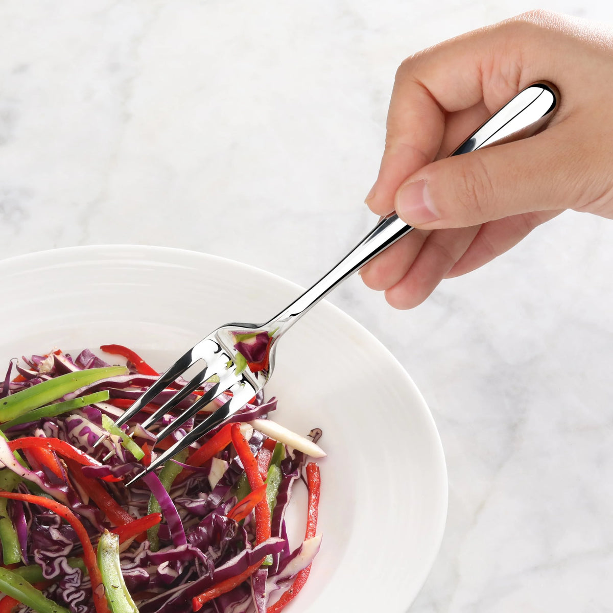 Hand holding a polished stainless steel salad fork spearing mixed sliced vegetables in a white bowl.