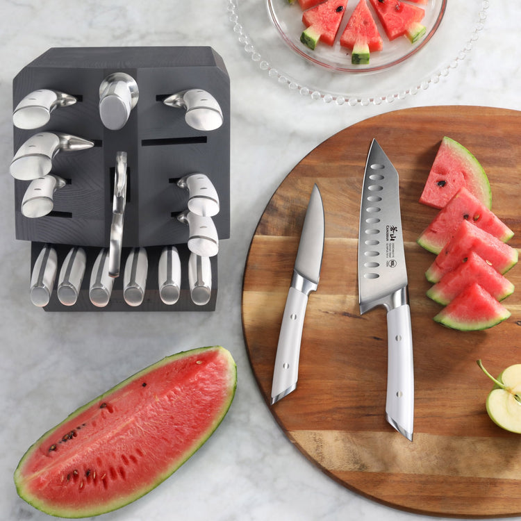 Black knife block with 17 stainless steel knives next to a wooden cutting board with sliced watermelon and two knives.