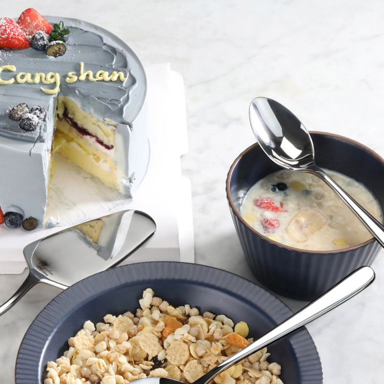 Serving spoon with mirror finish resting on a bowl of cereal, next to a cup of milk and a partially sliced cake.