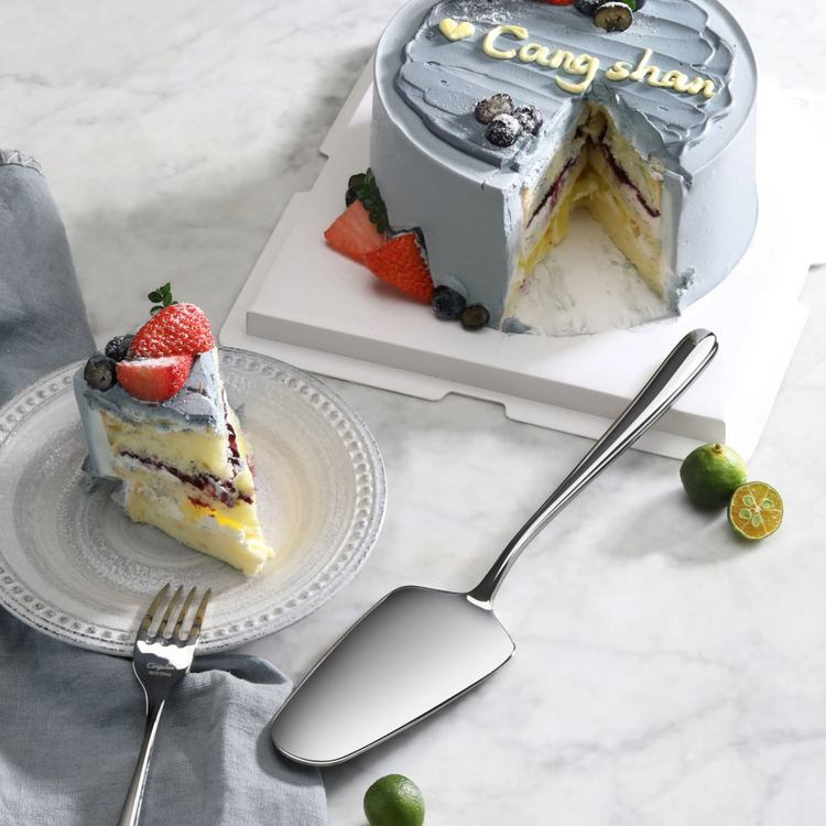 Polished stainless steel pie server next to a slice of layered cake with blueberries and strawberries on a plate.