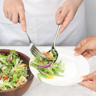 
                    
                      Two hands using a stainless steel serving fork and spoon to transfer salad from a wooden bowl to a white plate.
                    
                  
