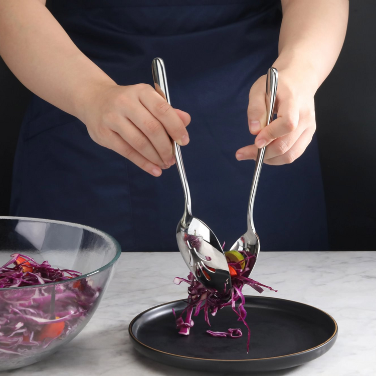 Person using two stainless steel slotted serving spoons to transfer shredded purple cabbage from a glass bowl to a black plat