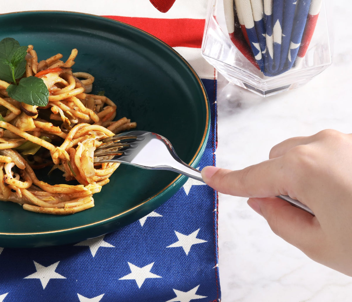 Hand holding stainless steel fork twirling spaghetti on dark green plate over star-patterned cloth.