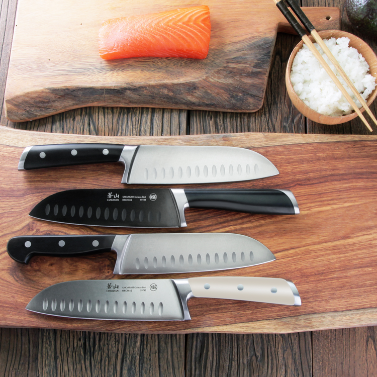 Four kitchen knives with black and white handles displayed on a wooden board, with a cutting board holding a piece of salmon