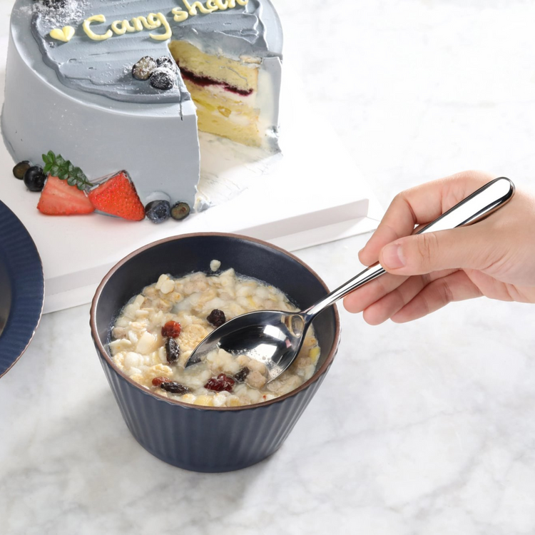 Hand holding a polished stainless steel serving spoon scooping oatmeal with dried fruit from a dark bowl, with a partially sl