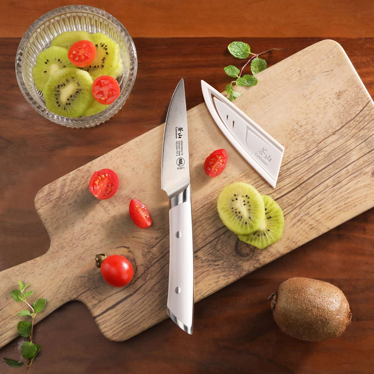 Paring knife with white handle and sheath on wooden cutting board with sliced kiwi and cherry tomatoes.