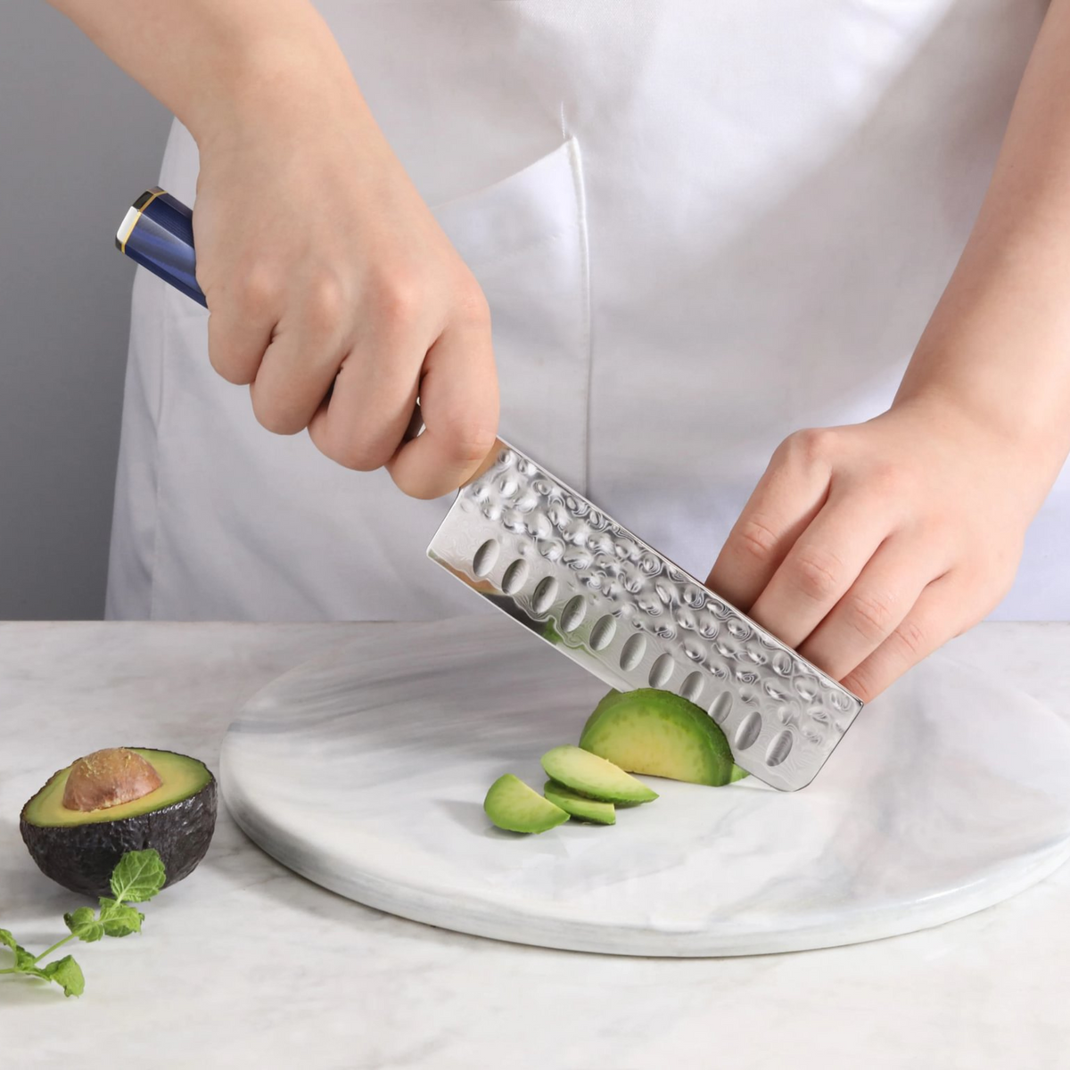 Hand holding a Nakiri knife slicing an avocado on a round marble cutting board.