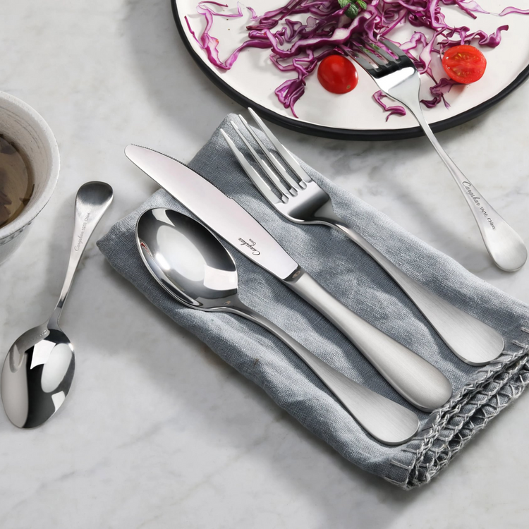Stainless steel flatware set with matte finish handles on a gray cloth, including a dinner spoon, knife, and fork.
