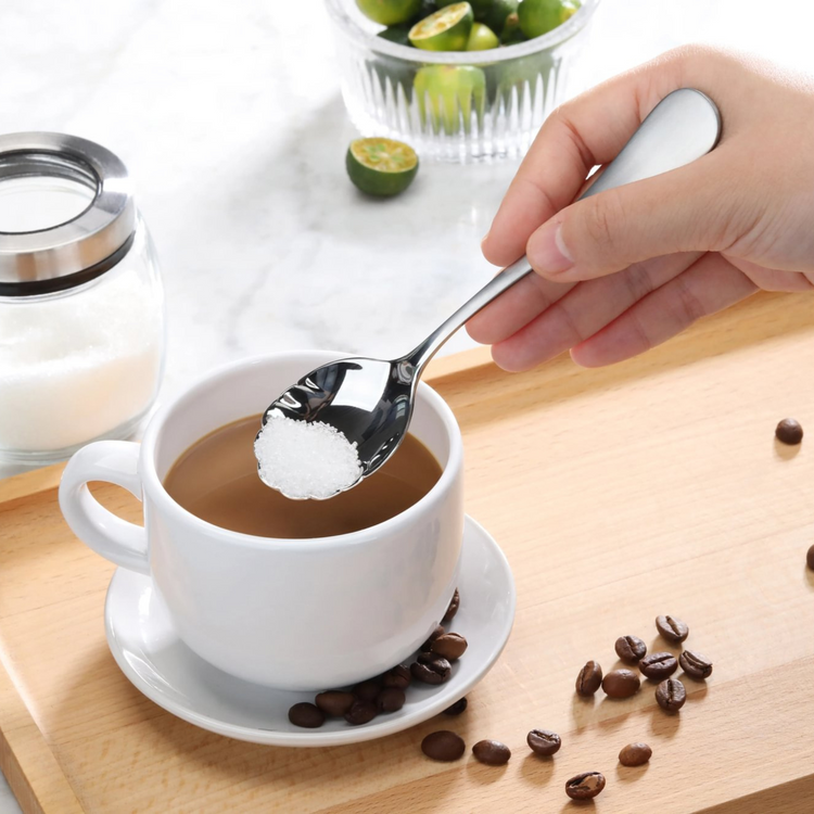 Hand holding a stainless steel sugar spoon with matte finish handles, scooping sugar over a cup of coffee.
