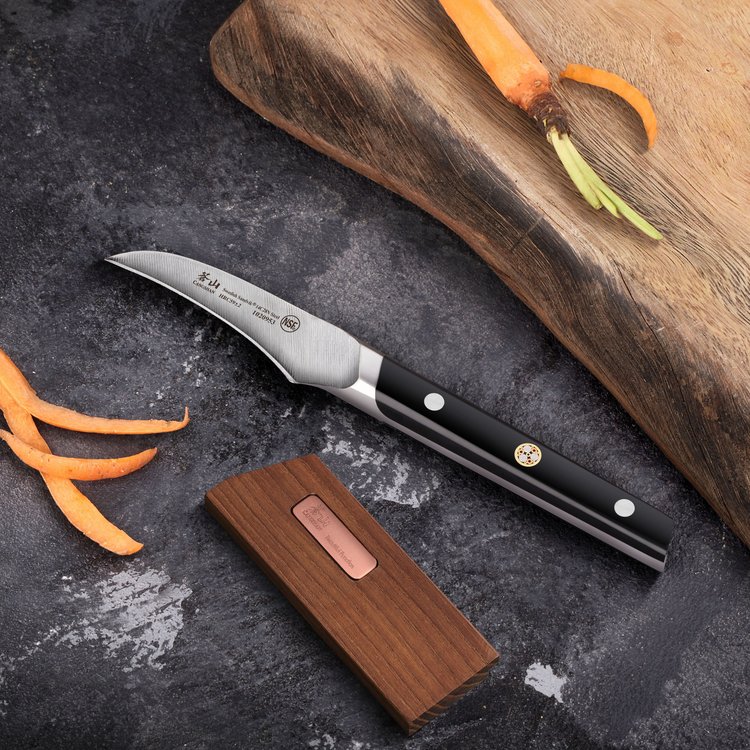 Paring knife with a curved blade, wooden sheath, and carrot peels on a dark countertop next to a wooden cutting board.