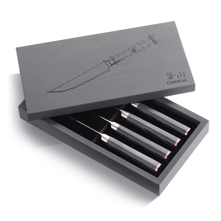 Four steak knives with black handles and silver blades in an open gray ash wood box with a knife sketch on the lid.