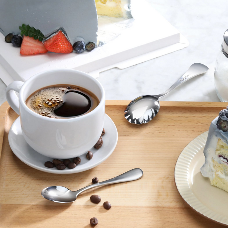 Stainless steel sugar spoon with matte finish handles on a wooden tray next to coffee cup and cake slices.