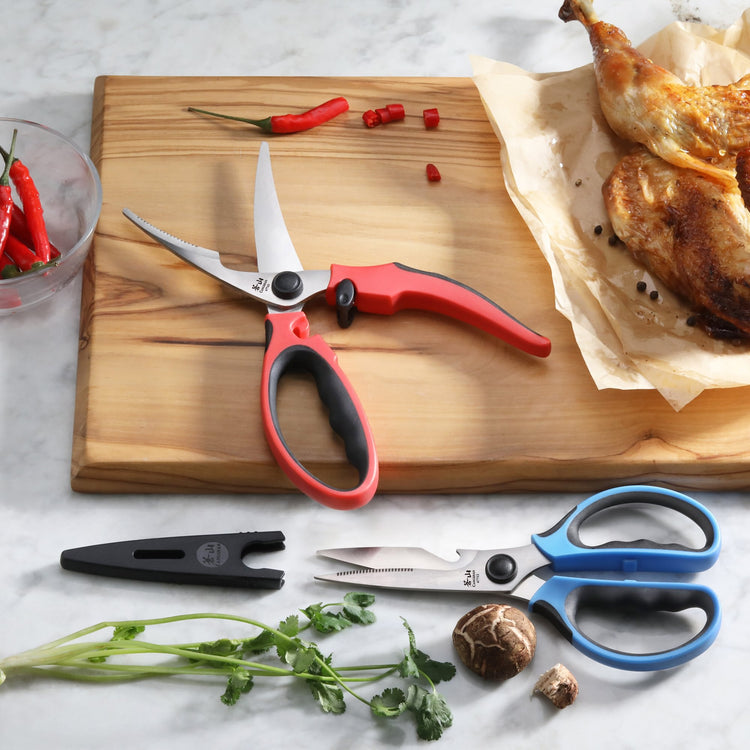 Two pairs of kitchen shears with colorful handles, one red and one blue, on a marble surface with herbs, mushrooms, and cooke