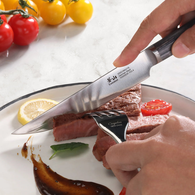 Hand using a steak knife with pakkawood handle to cut cooked steak on a plate with sauce, lemon slice, and cherry tomatoes.