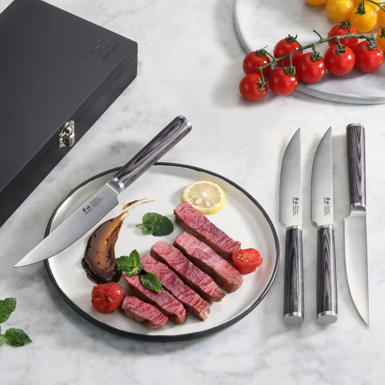 Four steak knives with pakkawood handles displayed next to a black wood box and a plate with sliced steak, cherry tomatoes, a