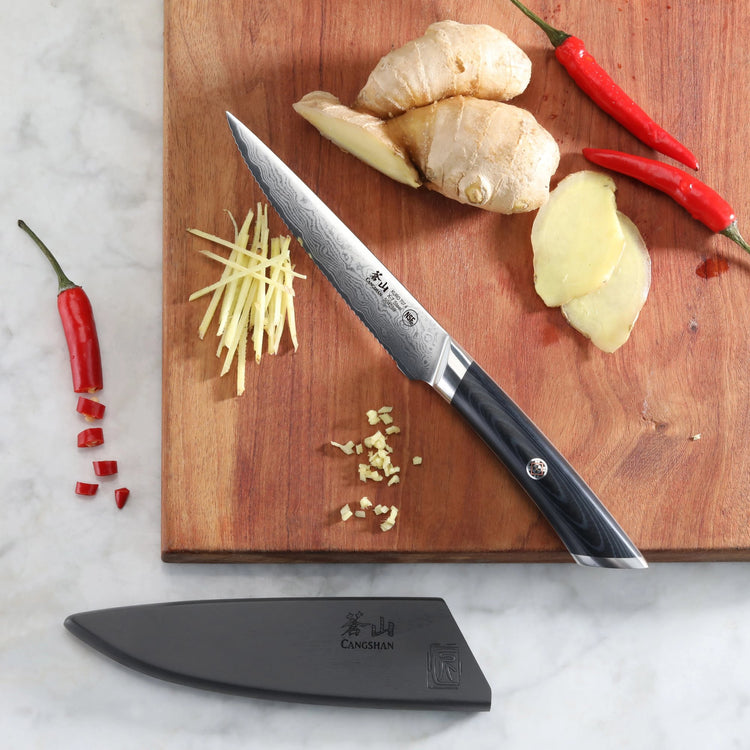 5-inch serrated utility knife with black handle on wooden board, sliced ginger and red chili peppers nearby, black sheath bel