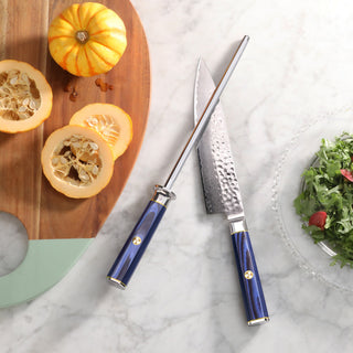 
                    
                      Blue-handled chef's knife and honing steel on marble surface next to wooden board with sliced squash and a bowl of greens.
                    
                  