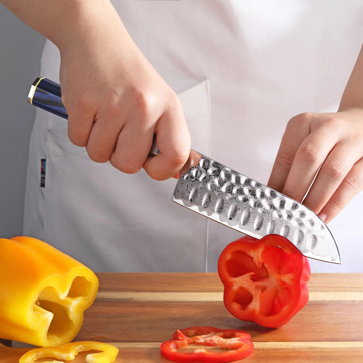 Hand holding a hammered steel Santoku knife slicing a red bell pepper on a wooden cutting board with a yellow bell pepper nea
