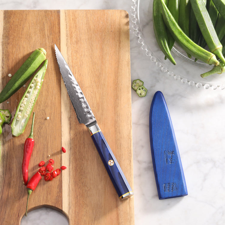 Serrated utility knife with blue handle and sheath on a wooden cutting board with sliced okra and red chili peppers.