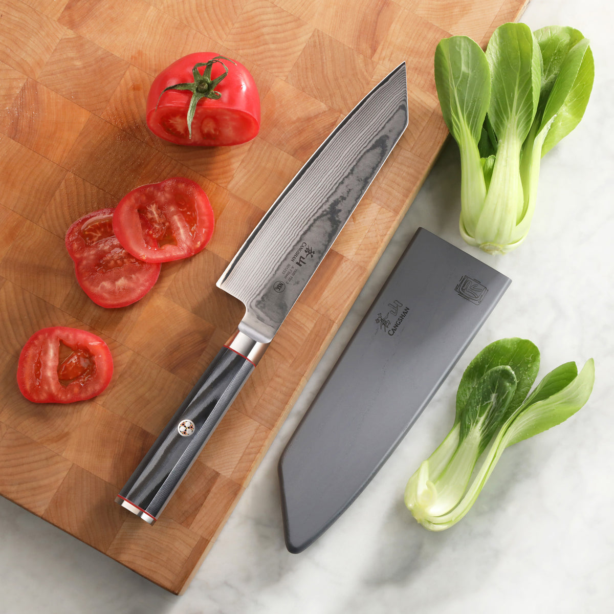 7-inch Kiritsuke knife with patterned blade and black handle, sheath, sliced tomato, and bok choy on cutting board and marble