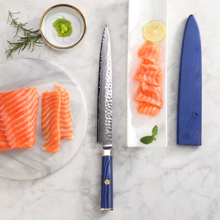 10-inch sashimi knife with blue handle and sheath, next to sliced salmon, lemon, wasabi, and herbs on marble surface.
