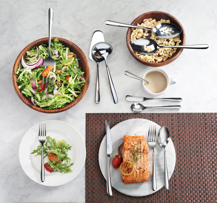 Two stainless steel spoons with mirror finish placed in wooden bowls with salad and cereal, alongside a table setting with sa