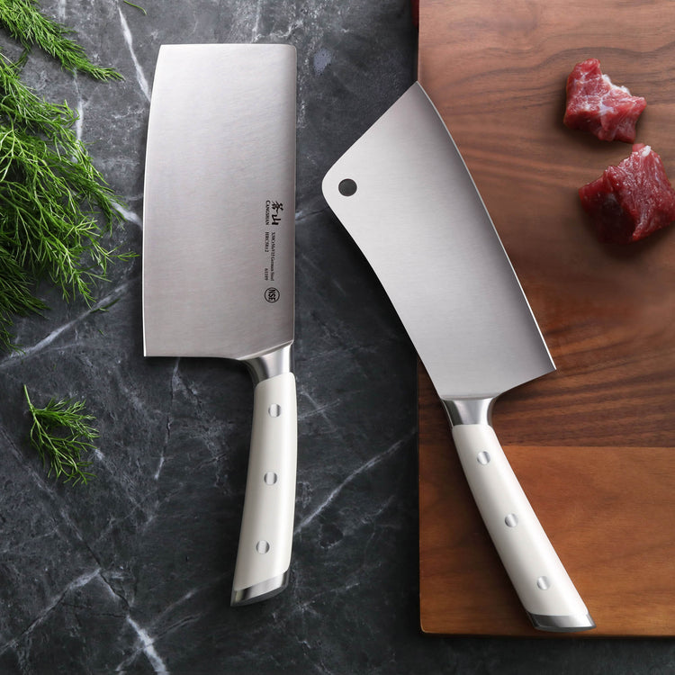Two stainless steel cleaver knives with white handles, one on a marble surface and one on a wooden cutting board with meat pi
