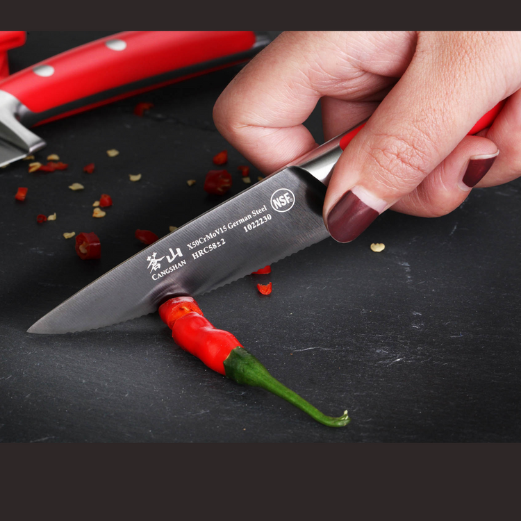 Hand with dark red nail polish slicing a red chili pepper on a black surface using a small serrated knife.