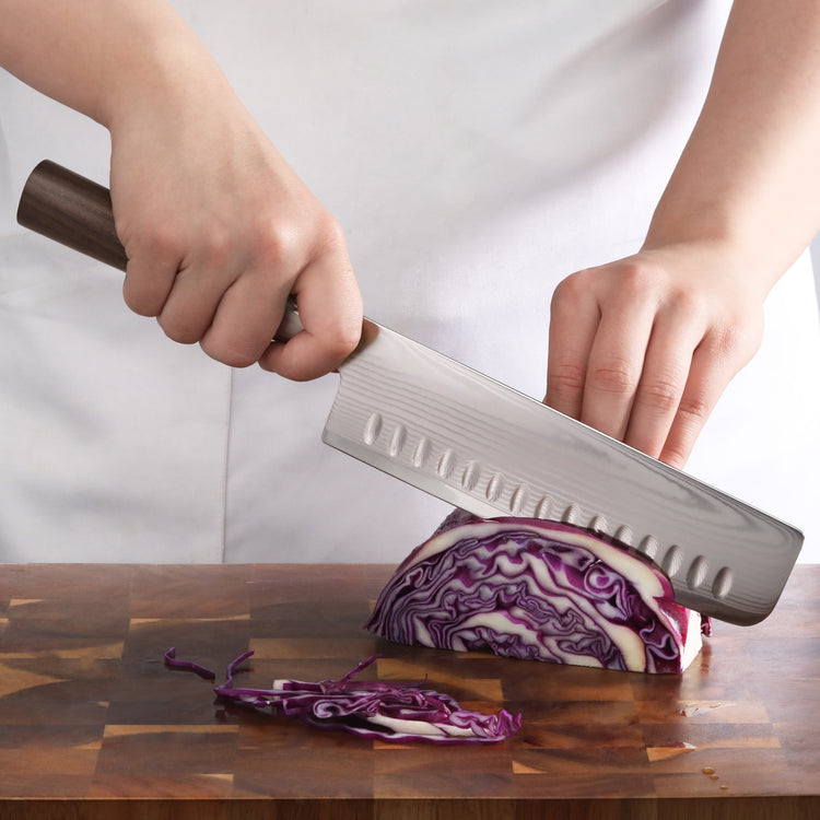 Hand holding a Nakiri knife slicing a wedge of purple cabbage on a wooden cutting board.