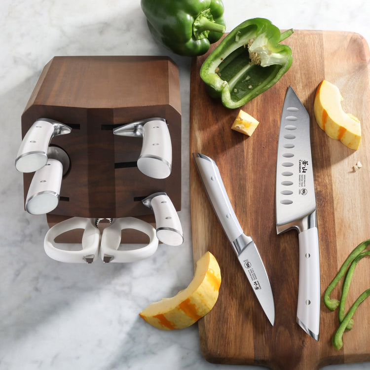 Wooden knife block with six knives and kitchen shears next to a cutting board holding two knives, a green bell pepper, and pu