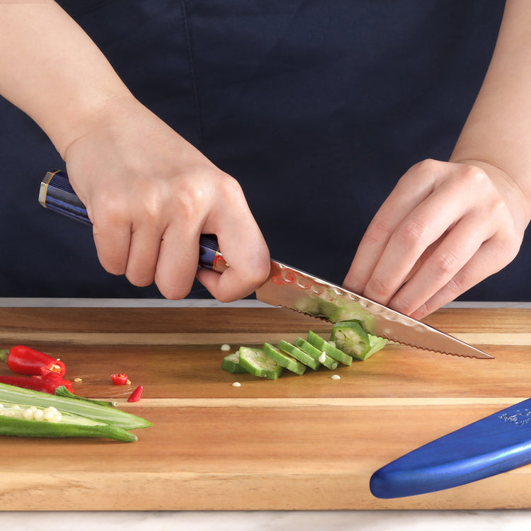 Hands slicing a green vegetable with a serrated knife on a wooden cutting board, blue sheath nearby.