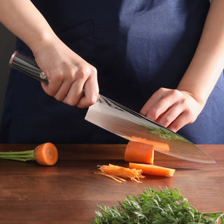 
                    
                      Hands holding a chef's knife slicing a carrot on a wooden cutting board with carrot greens nearby.
                    
                  
