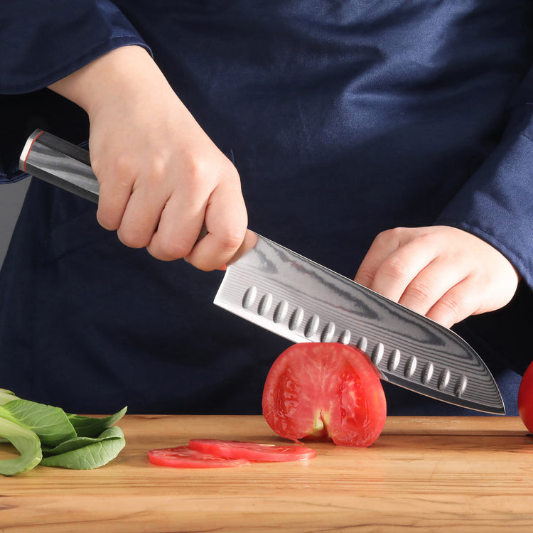Hand holding YARI Series 7-Inch Santoku Knife slicing a tomato on a wooden cutting board.
