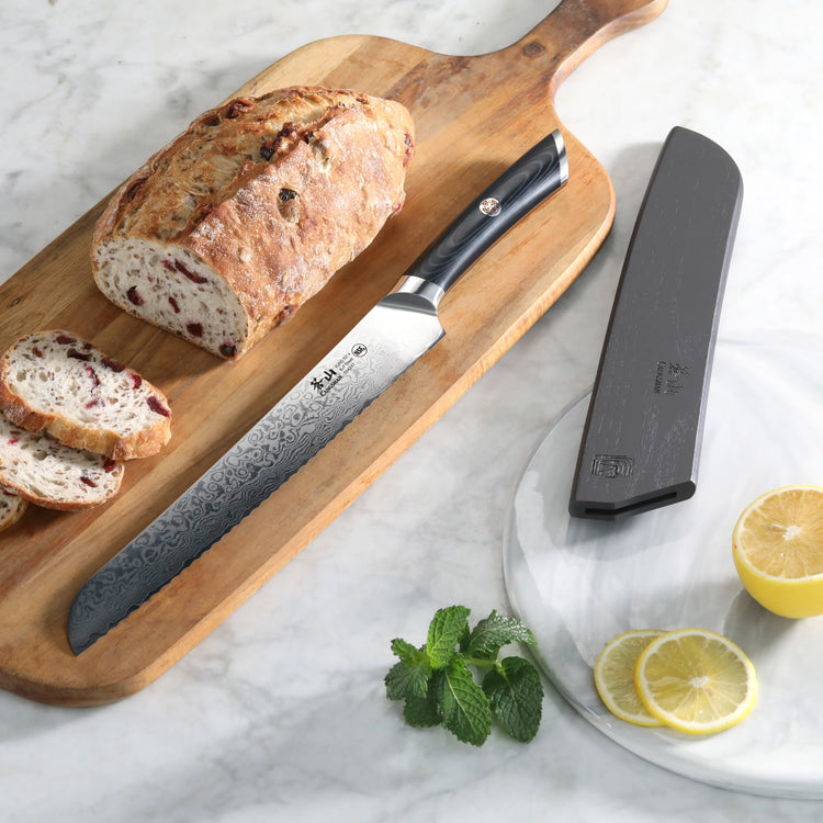 Bread knife with patterned blade and black handle on wooden board next to sliced bread, knife sheath, lemon slices, and mint