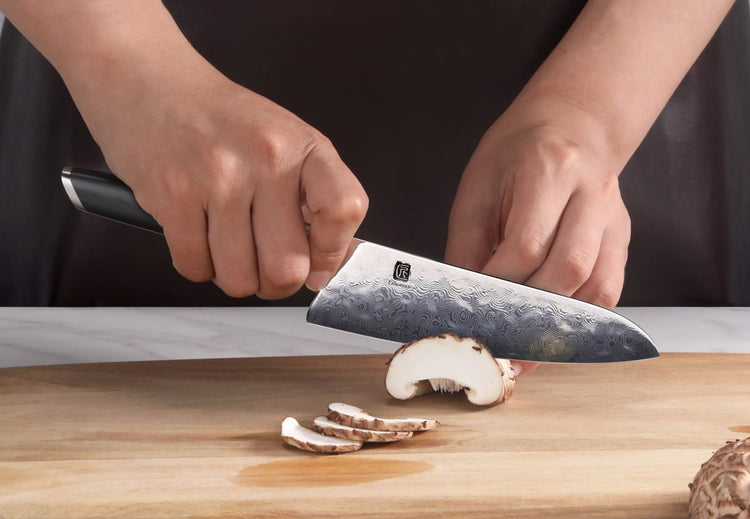 Hands slicing a mushroom on a wooden cutting board with a patterned steel utility knife.