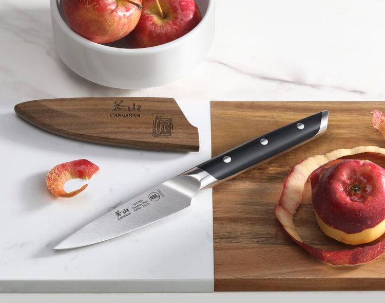 3.5-inch paring knife with black handle and wooden sheath next to peeled red apple on cutting board.