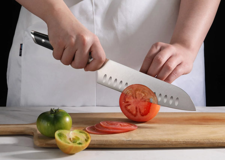 Person slicing a red tomato on a wooden cutting board with a Santoku knife, next to green and yellow tomatoes.