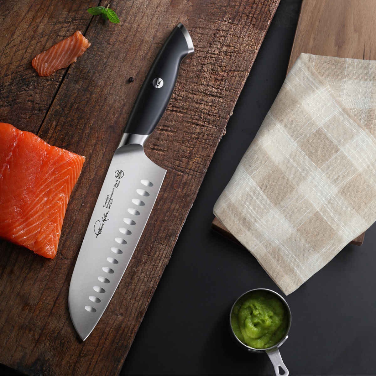 Santoku knife with black handle on wooden board next to salmon pieces, beige cloth, and green sauce in measuring cup.