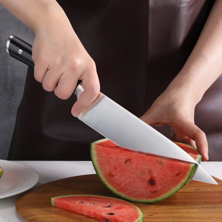 Hand holding a chef knife slicing a watermelon wedge on a wooden cutting board.