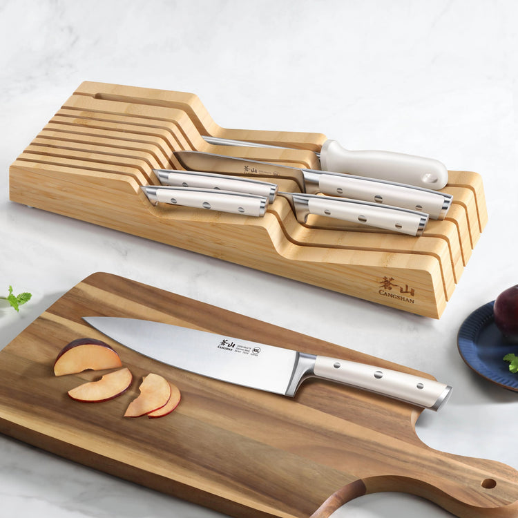 Seven knives with stainless steel blades and white handles in a wooden in-drawer block, one knife slicing apple on a wooden c