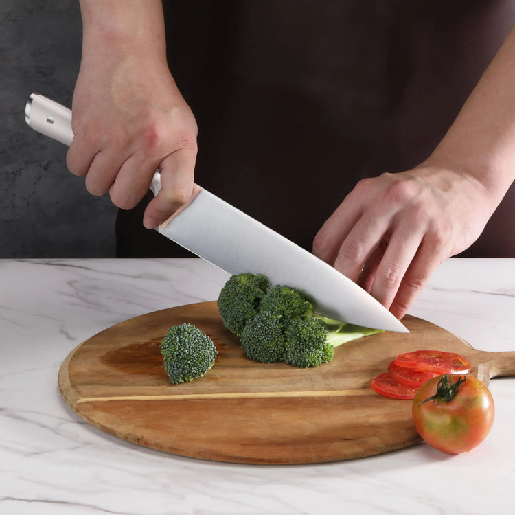 Hands slicing broccoli on a wooden cutting board with a large kitchen knife, next to sliced tomatoes and a whole tomato.