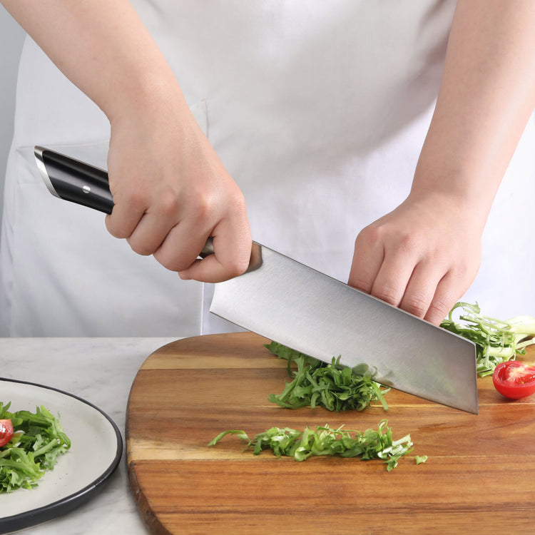 Person chopping green herbs on a wooden cutting board with a black-handled Nakiri knife.