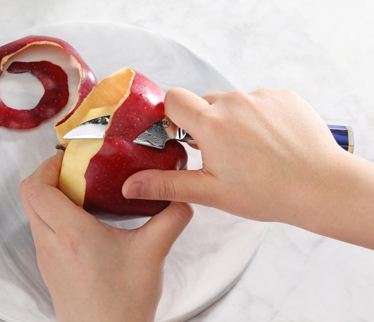 Hands peeling a red apple with a small blue-handled knife on a white plate.
