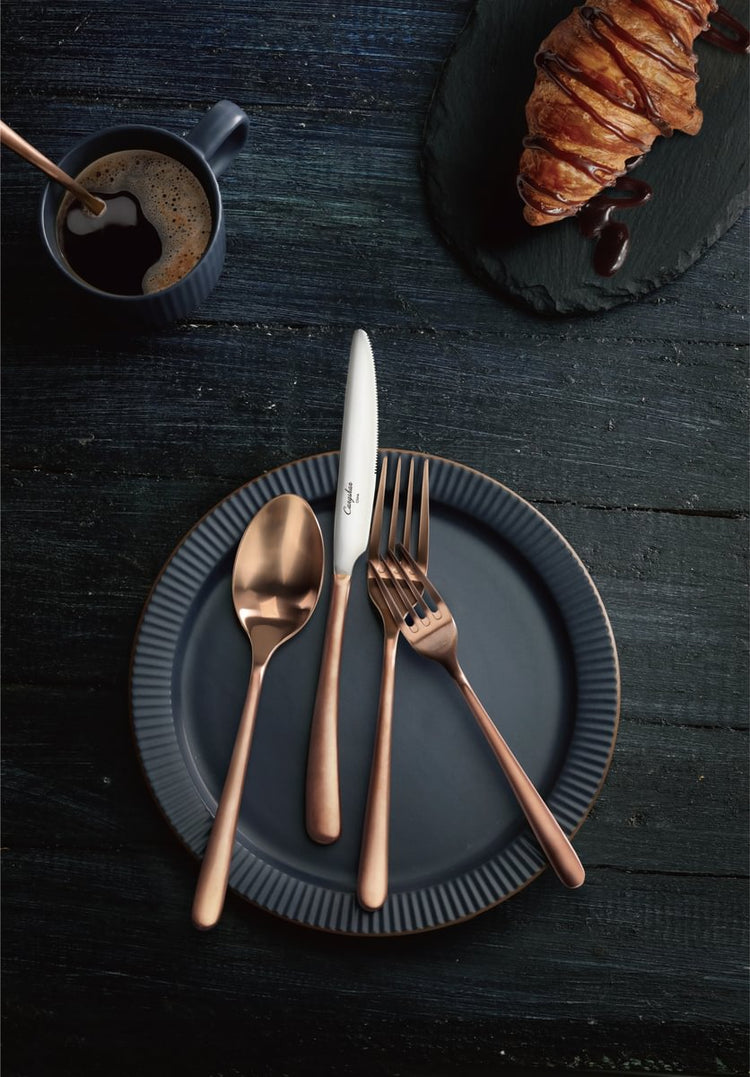 Copper flatware set with a spoon, knife, and two forks on a dark plate next to a cup of coffee and a croissant.
