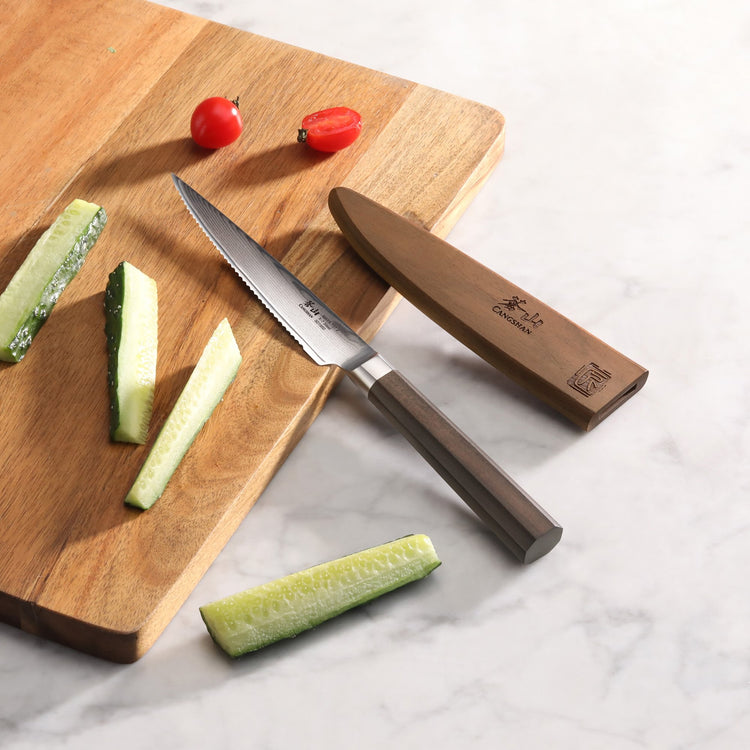 MAYA Series 5-inch serrated utility knife with sheath on a cutting board near sliced cucumbers and tomatoes.